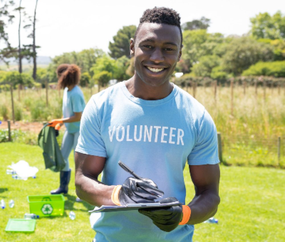 Portrait of a smiling young African American man standing in a field wearing wearing a t shirt with volunteer written on it and gloves, writing on a clipboard and smiling to camera, while a diverse group of volunteers collect rubbish and recycling in the background