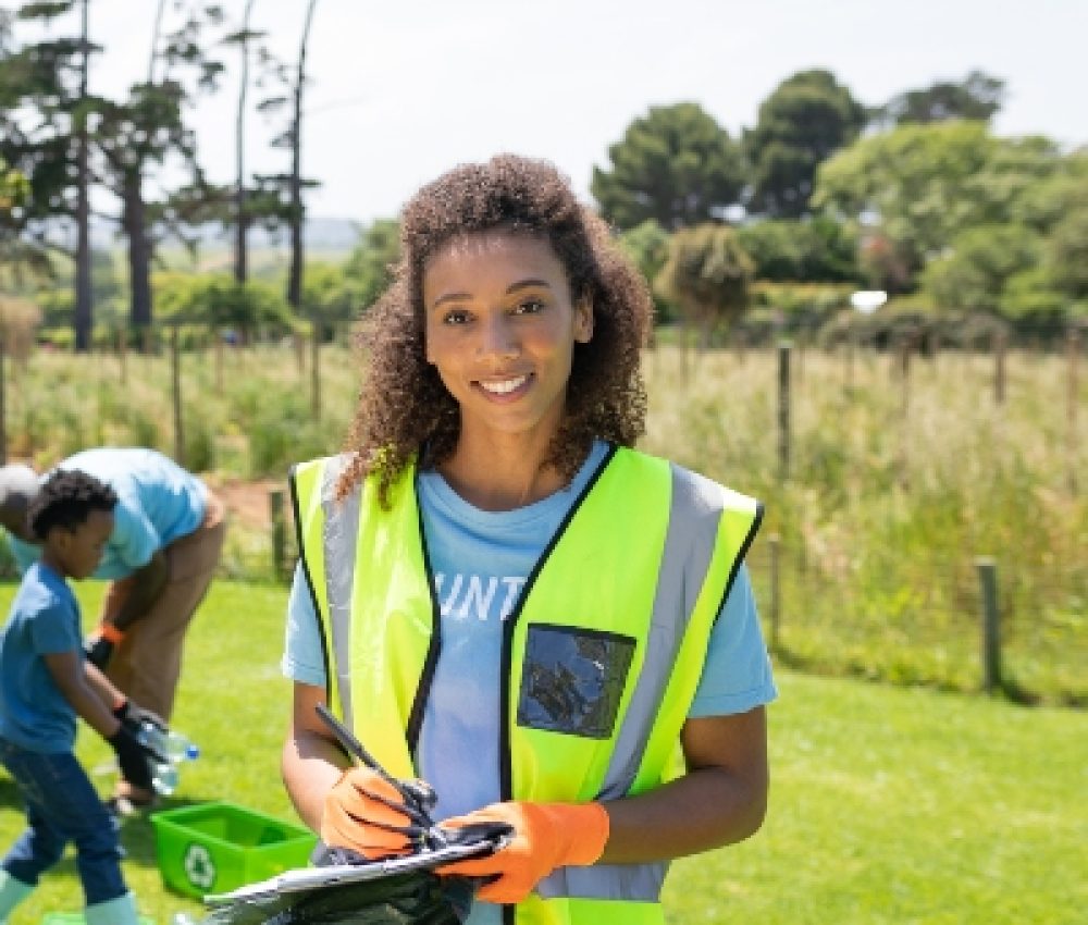 Front view of a young African American woman standing in a field wearing a hi-vis vest and gloves, writing on a clipboard and smiling to camera, while a diverse group of volunteers collect rubbish and recycling in the background